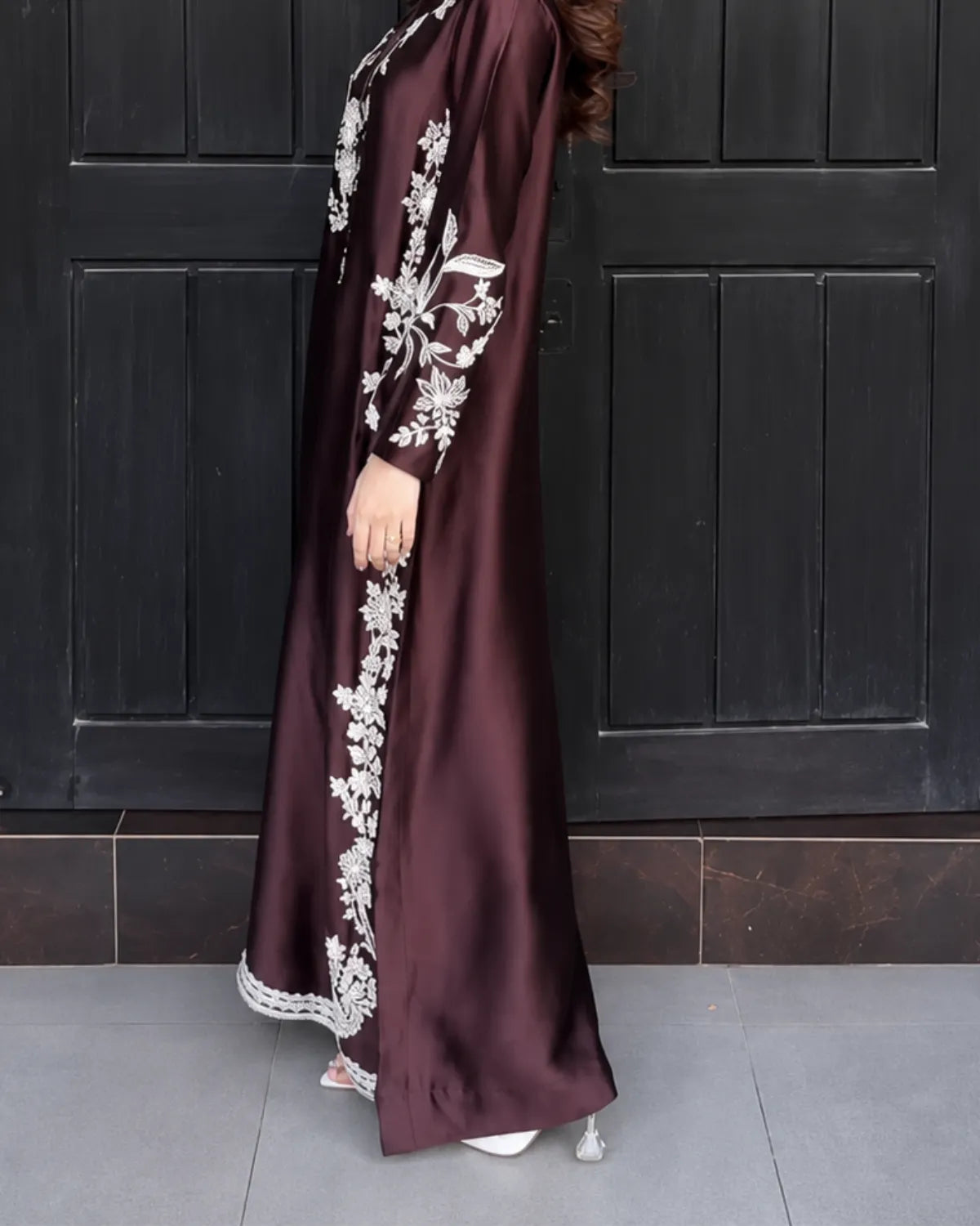 A woman wearing a long, flowing dark plum abaya with intricate white floral embroidery and lace trim, standing in front of a dark paneled door.