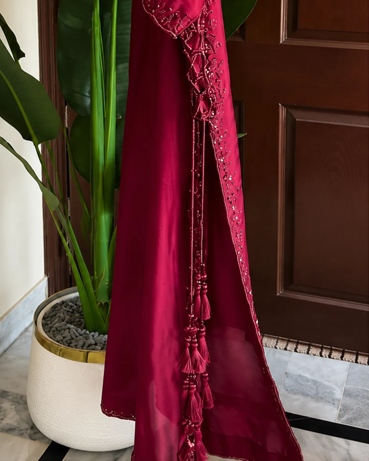 A deep magenta-red satin dress or caftan with intricate beadwork and tassel details, draped next to a potted plant in front of a dark wooden door.