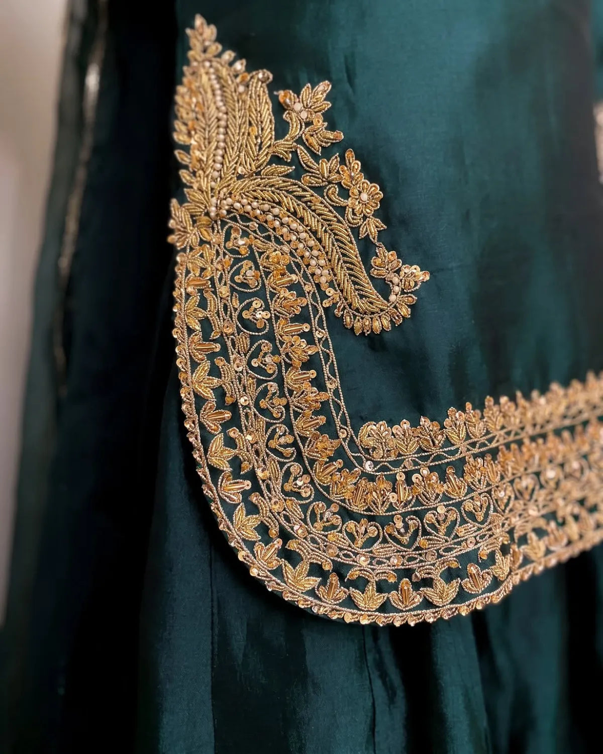 Close-up of a dark teal green fabric garment featuring an elaborate, wide gold embroidered border with intricate floral and paisley patterns.