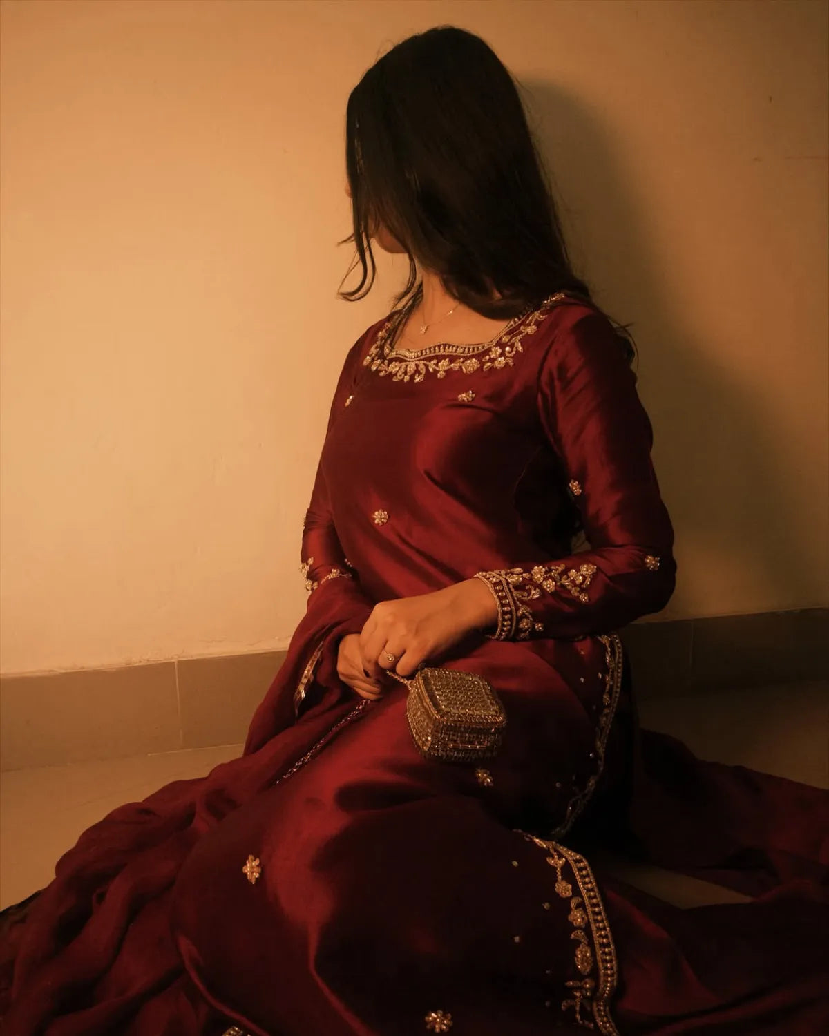 A woman in a deep maroon traditional outfit with gold embroidery sits against a plain beige wall, holding a small metallic clutch bag.