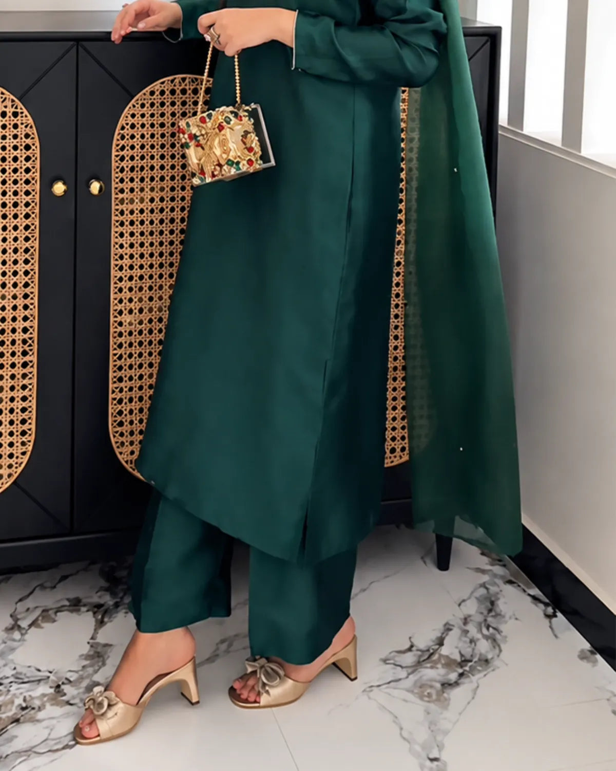 A person wearing a dark green traditional outfit with matching pants and a sheer dupatta, holding a small floral handbag, standing indoors near a black cabinet with woven panels.