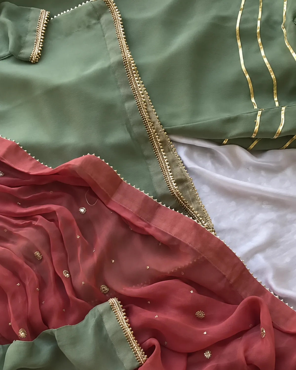 A close-up of a traditional Indian outfit featuring a sage green kurta with gold zari embroidery, a dusty pink dupatta with gold motifs, and a white undergarment.