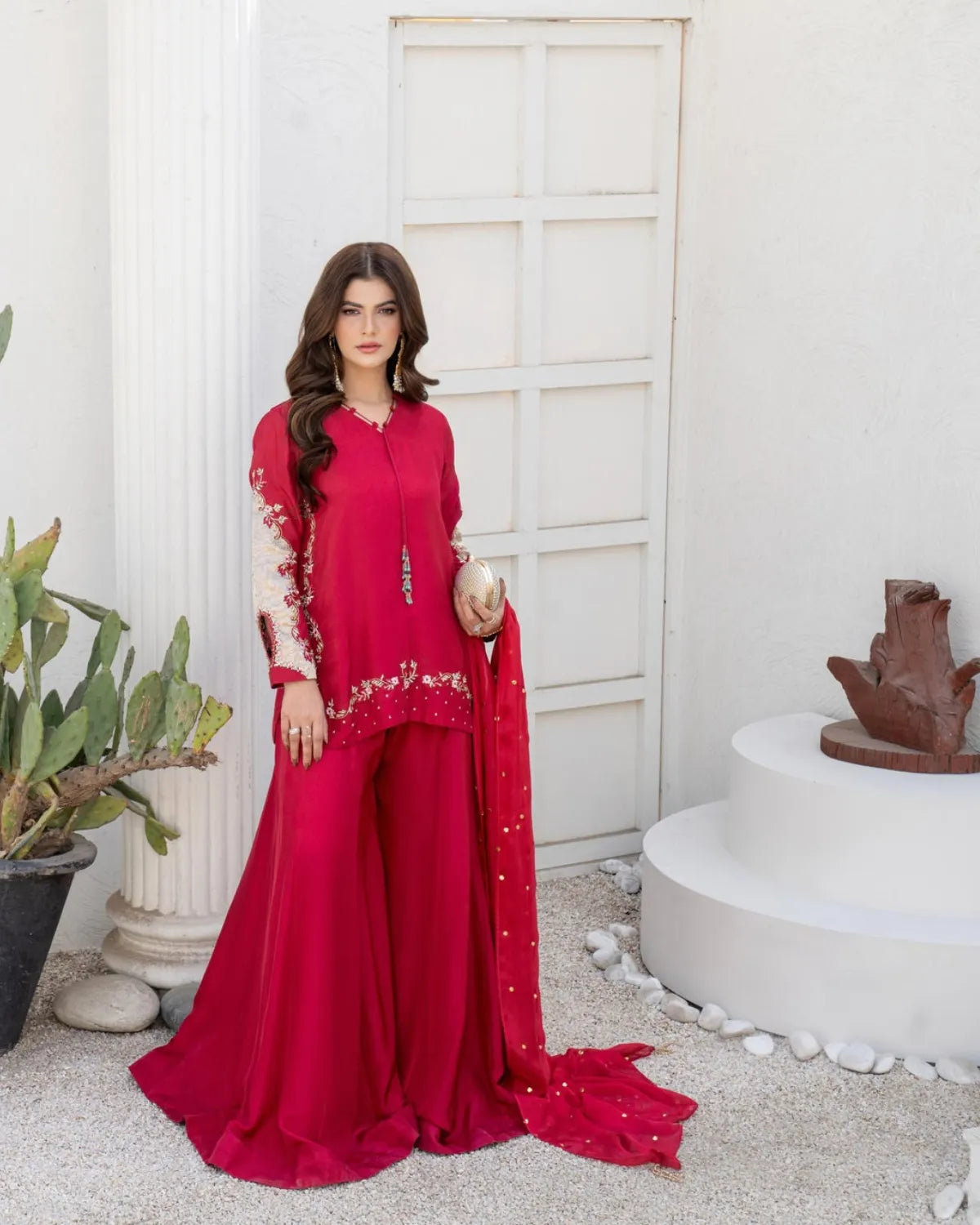 A woman models a vibrant red three-piece traditional outfit with intricate gold embroidery, standing in front of a white stucco wall with decorative plants and a sculpture.