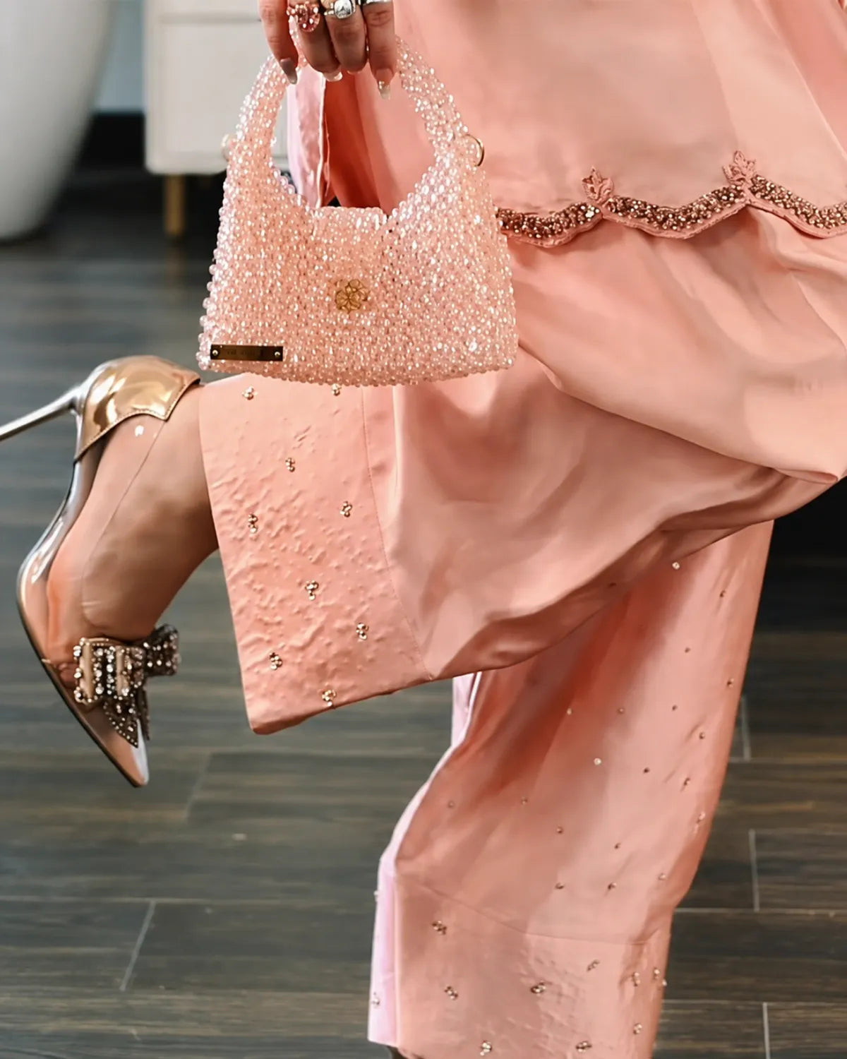 A close-up lifestyle shot of a person wearing a peach pink embroidered traditional outfit, holding a matching beaded handbag and wearing metallic rose gold pointed-toe heels with jeweled embellishments on the toes.