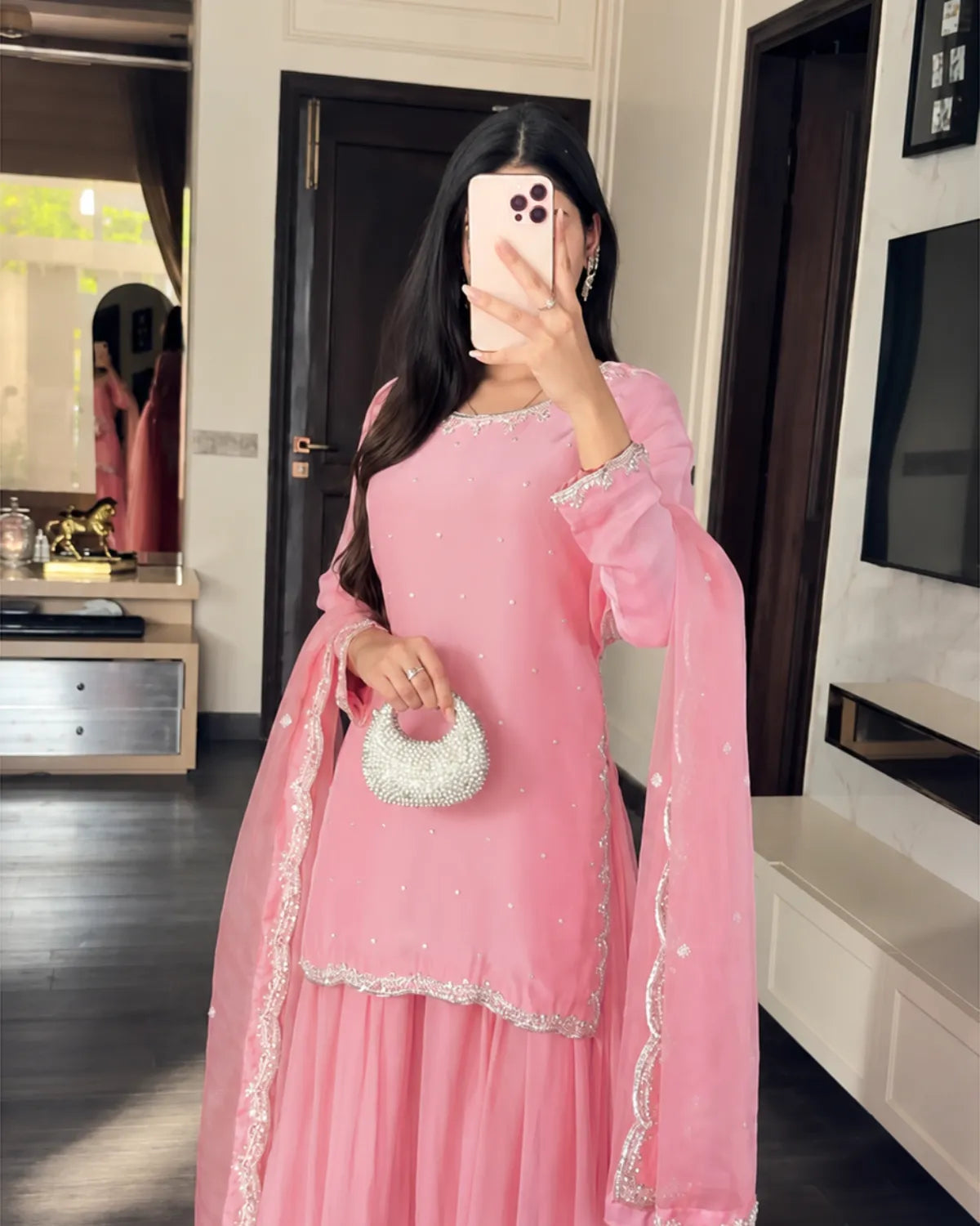 A woman takes a mirror selfie wearing a pink traditional outfit with silver embroidery and a matching dupatta, holding a small white beaded handbag in a modern indoor setting.