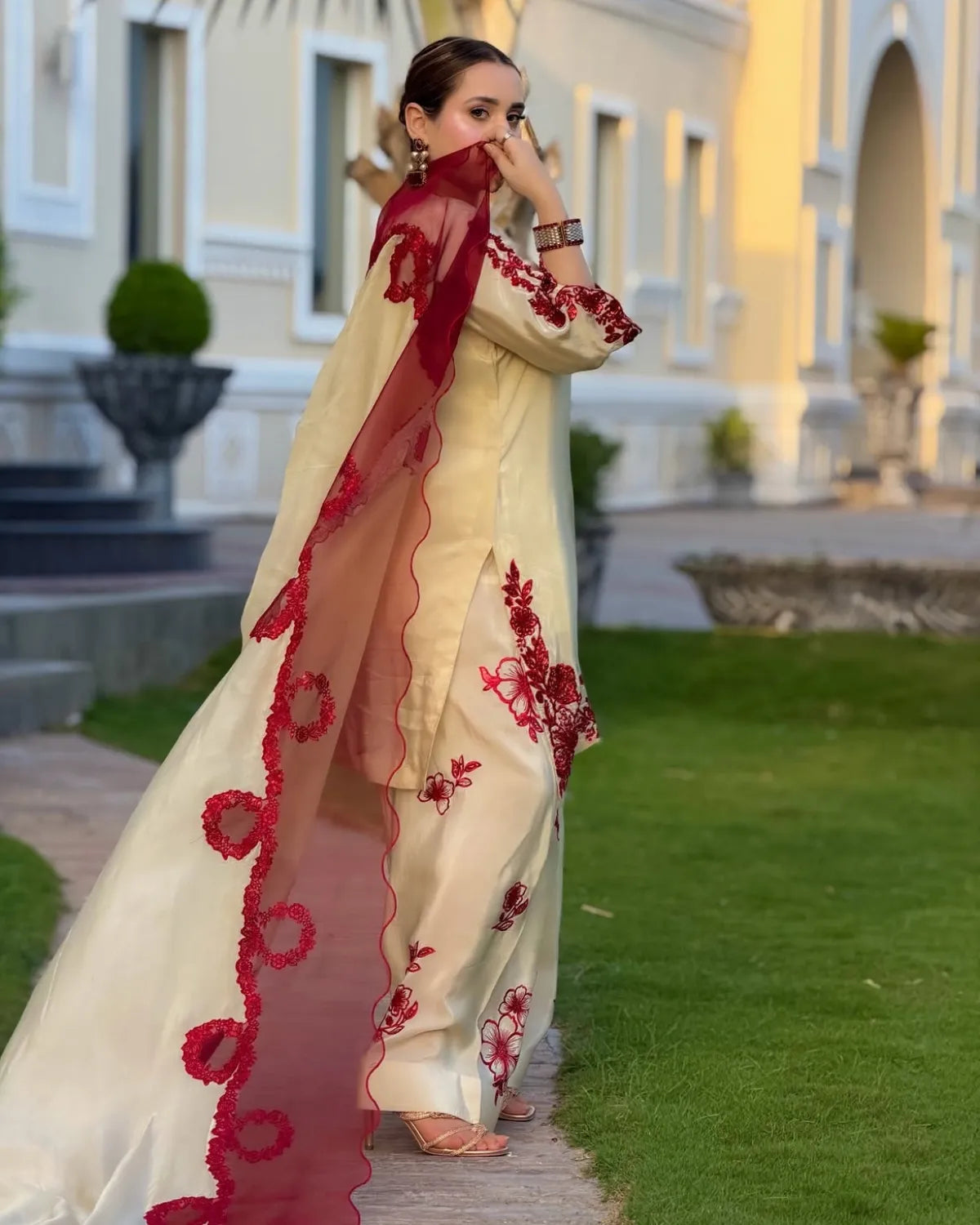 A woman wearing a cream-colored traditional outfit with red floral embroidery and a matching sheer red dupatta, standing outdoors on a garden path with a building in the background.