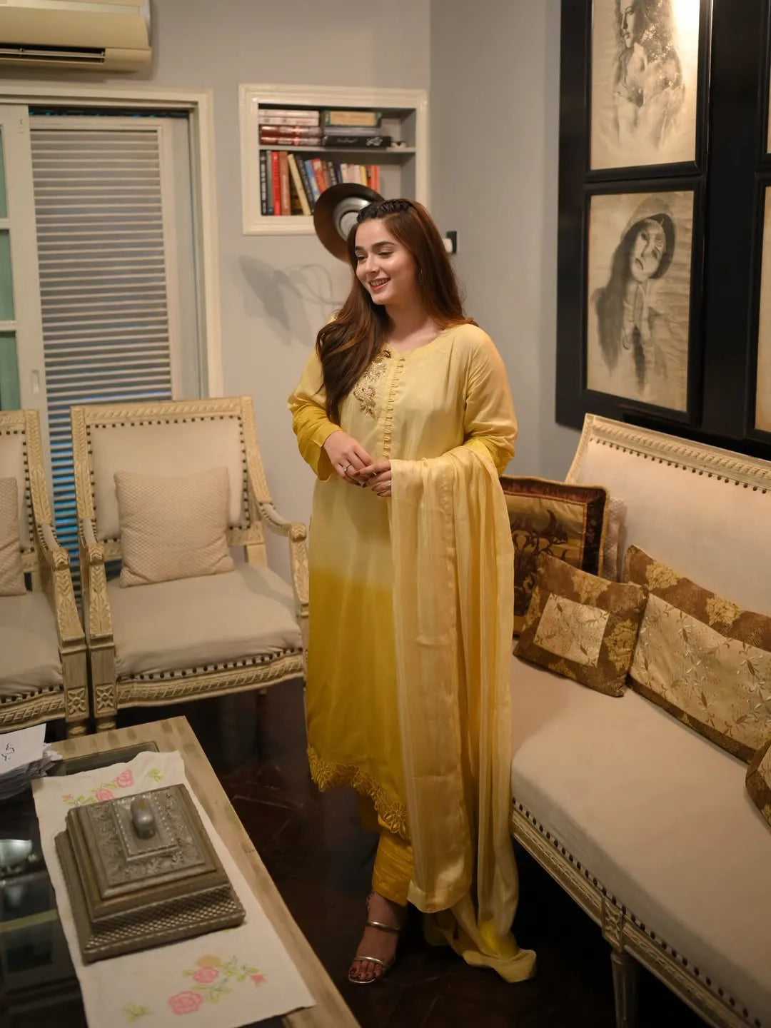 Woman in a yellow traditional outfit standing in a living room with decor elements.