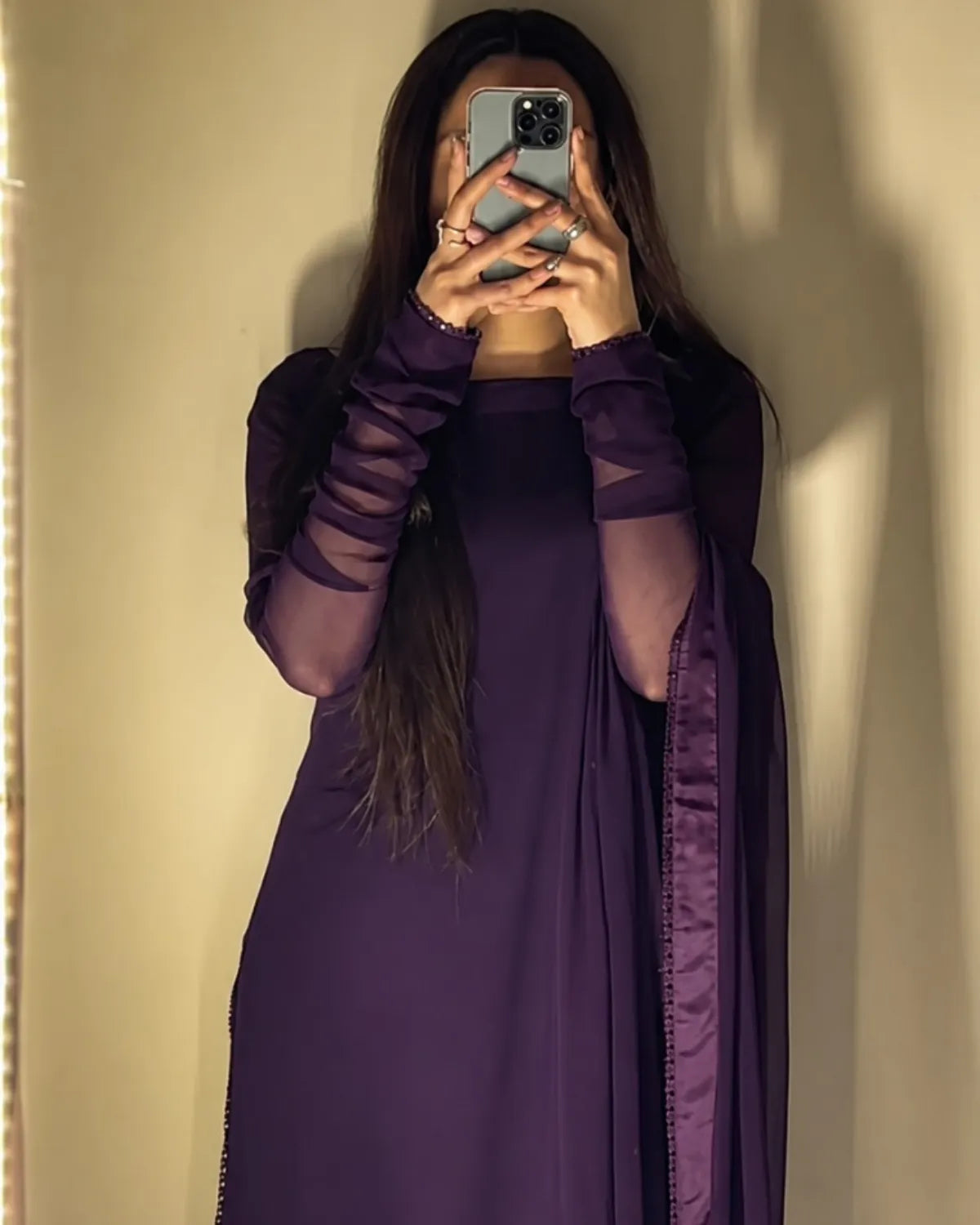 Person wearing a long purple dress with sheer sleeves against a neutral wall.