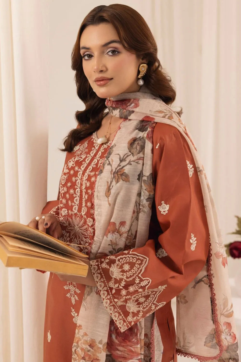 Woman wearing a traditional outfit with a floral shawl, holding a book against a white curtain background.