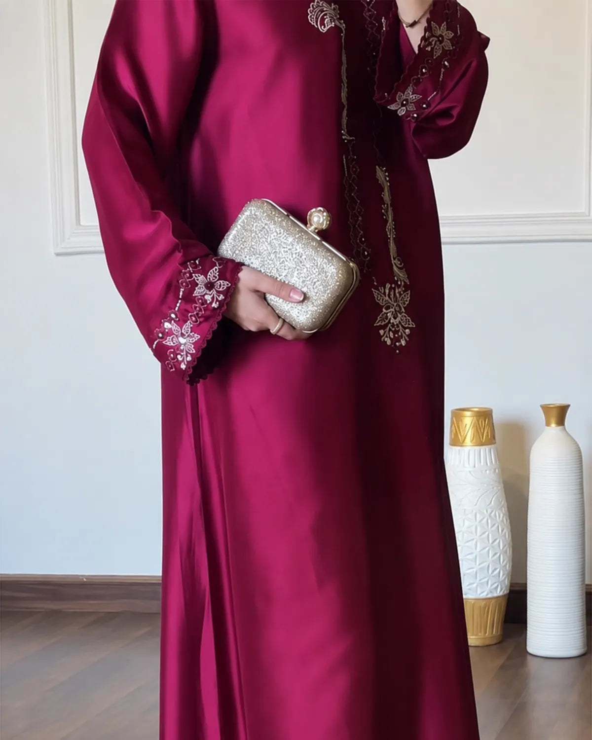 A person wearing a long magenta satin abaya dress with gold floral embroidery on the sleeves and chest, holding a glittering gold clutch purse in an indoor setting.