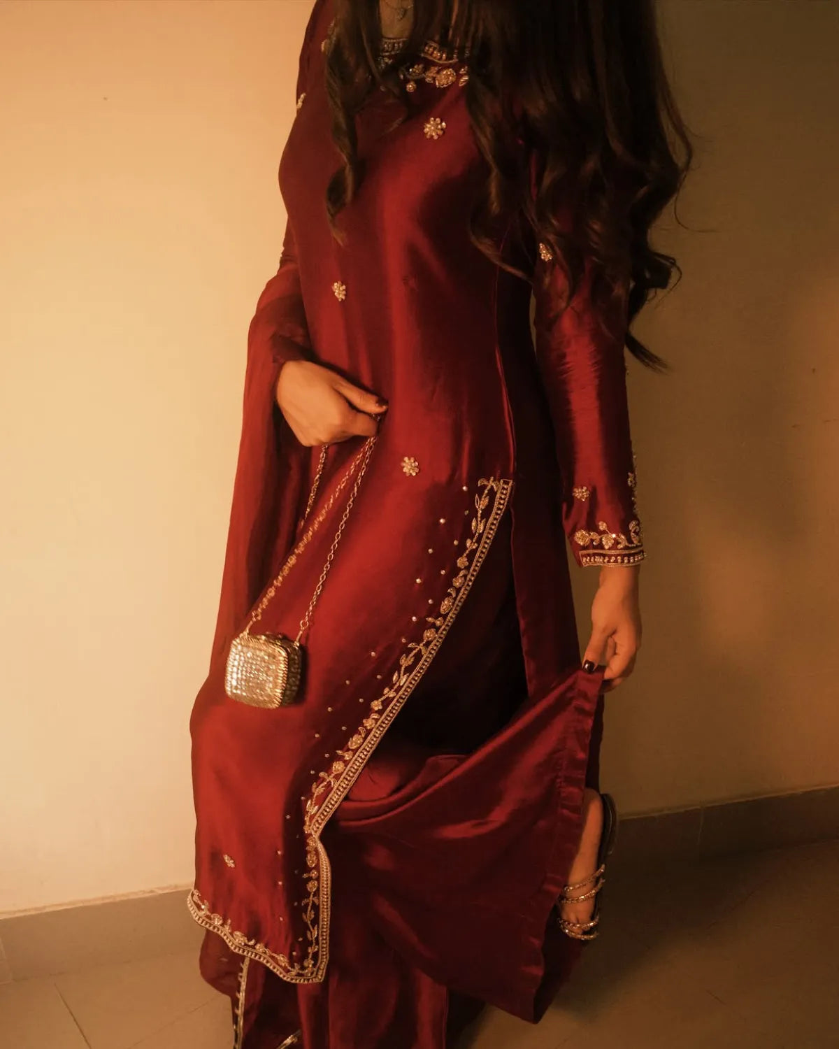 A woman wearing a deep maroon red satin Anarkali kurta with gold embroidery, holding a matching embroidered dupatta and a small silver purse.