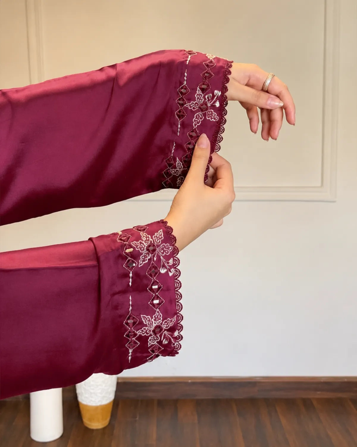 A close-up of a person's hands and arms wearing a long-sleeved burgundy satin top with intricate white floral embroidery and scalloped lace cuffs, set against an indoor wall background.