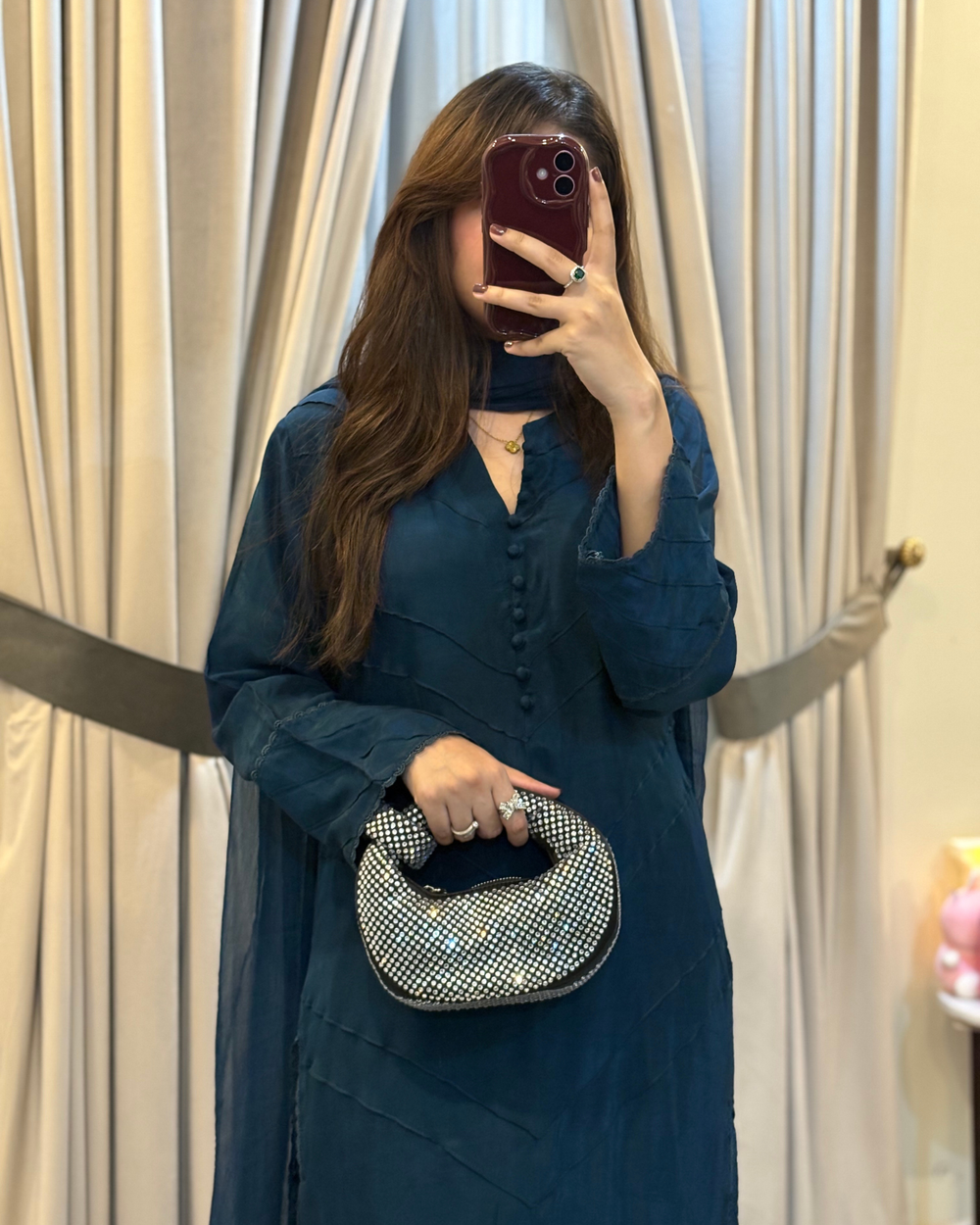 Woman in a teal dress holding a silver clutch in front of beige curtains