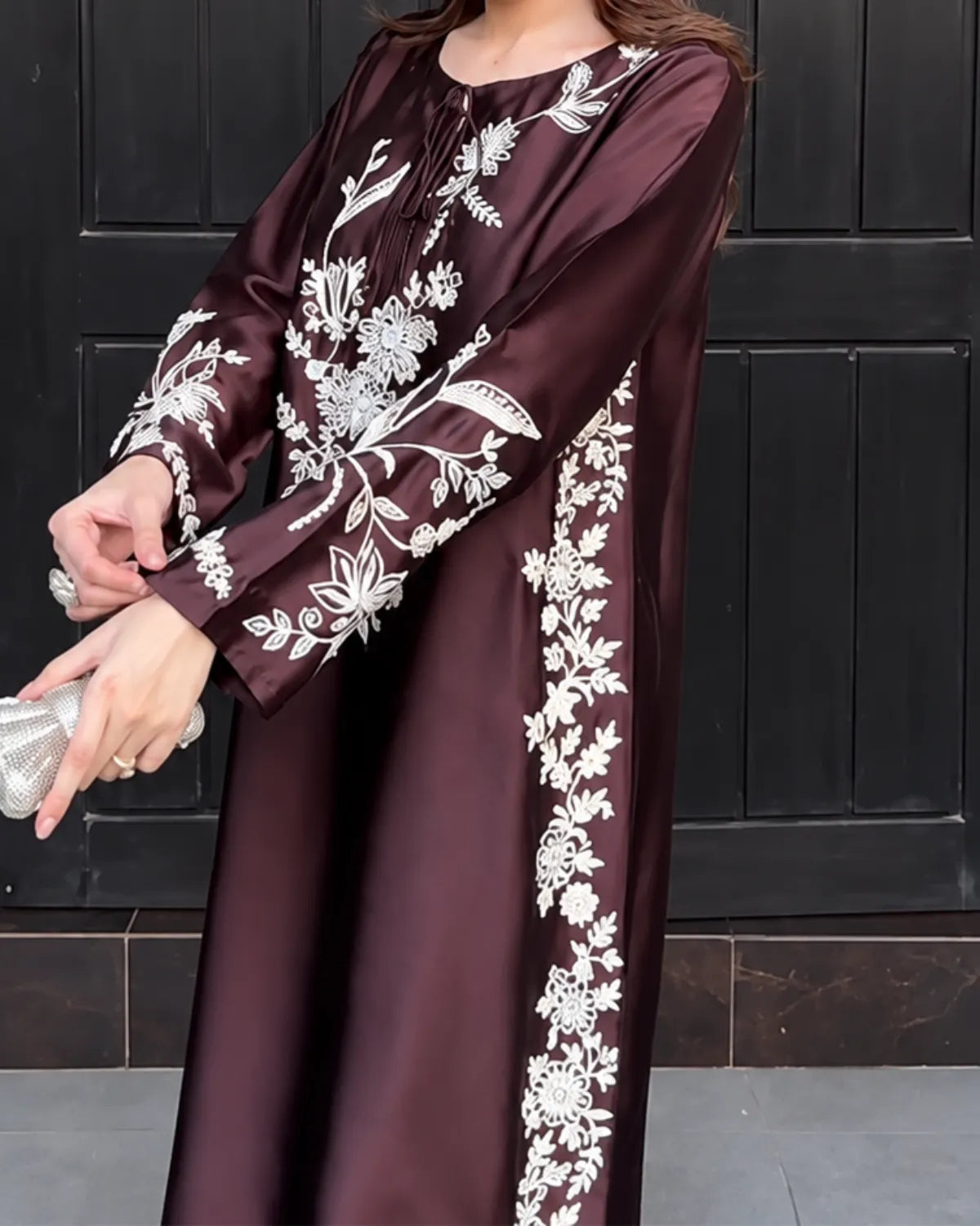 A woman wearing a long-sleeved, floor-length abaya dress in deep burgundy with intricate white floral embroidery, holding a silver clutch in front of a dark paneled door.