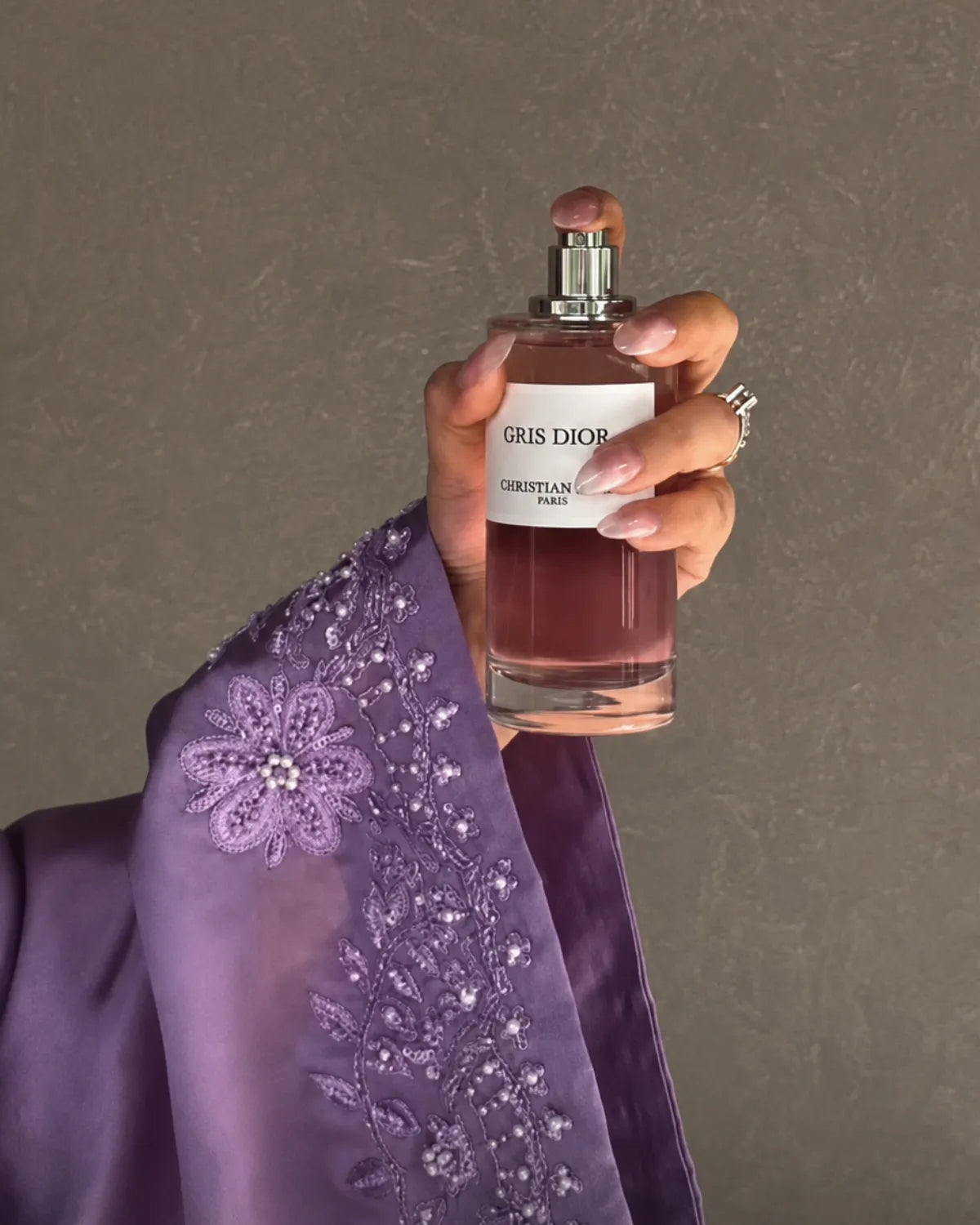 A hand holding a bottle of Dior Gris Dior perfume, wearing a purple embroidered sleeve against a textured gray background.