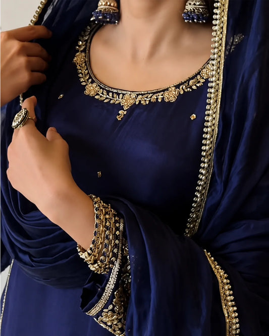 A close-up of a navy blue embroidered traditional outfit with ornate gold and silver embellishments, worn by a model. The outfit features a deep neckline decorated with detailed metallic embroidery, a matching embroidered border on the draped fabric, and coordinating jewelry including earrings and a ring.