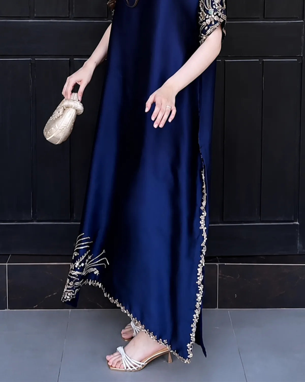 A woman wearing a long navy blue satin dress with gold and silver embroidered details on the sleeves and hem, holding a small gold clutch against a dark paneled background.