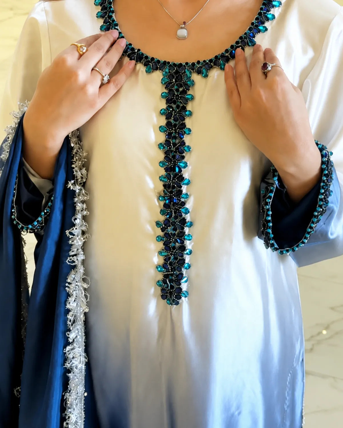 A close-up of a person wearing a cream satin tunic with a deep V-neckline and sleeve cuffs decorated with dark blue and turquoise beadwork, along with a matching blue draped shawl.