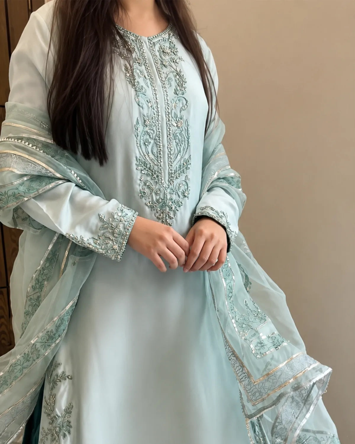 A woman wearing a light blue-green traditional South Asian outfit with intricate silver embroidery on the neckline, sleeves, and dupatta, set against a plain beige wall.