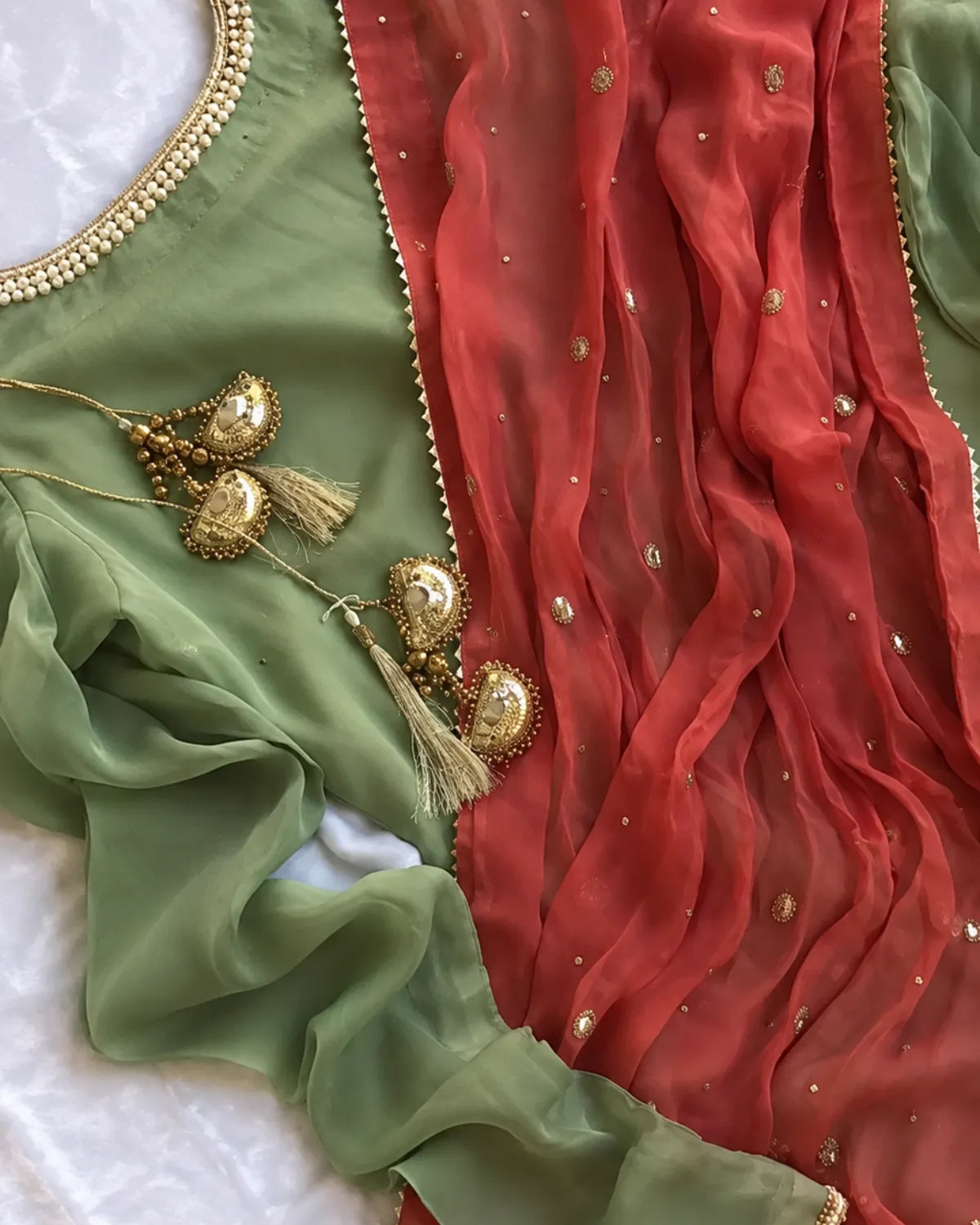 A close-up of a traditional Indian ethnic outfit fabric set featuring a sage green blouse with gold embroidery, a rust red sheer dupatta with gold zari work, and ornate gold tassel jewelry laid on a white surface.