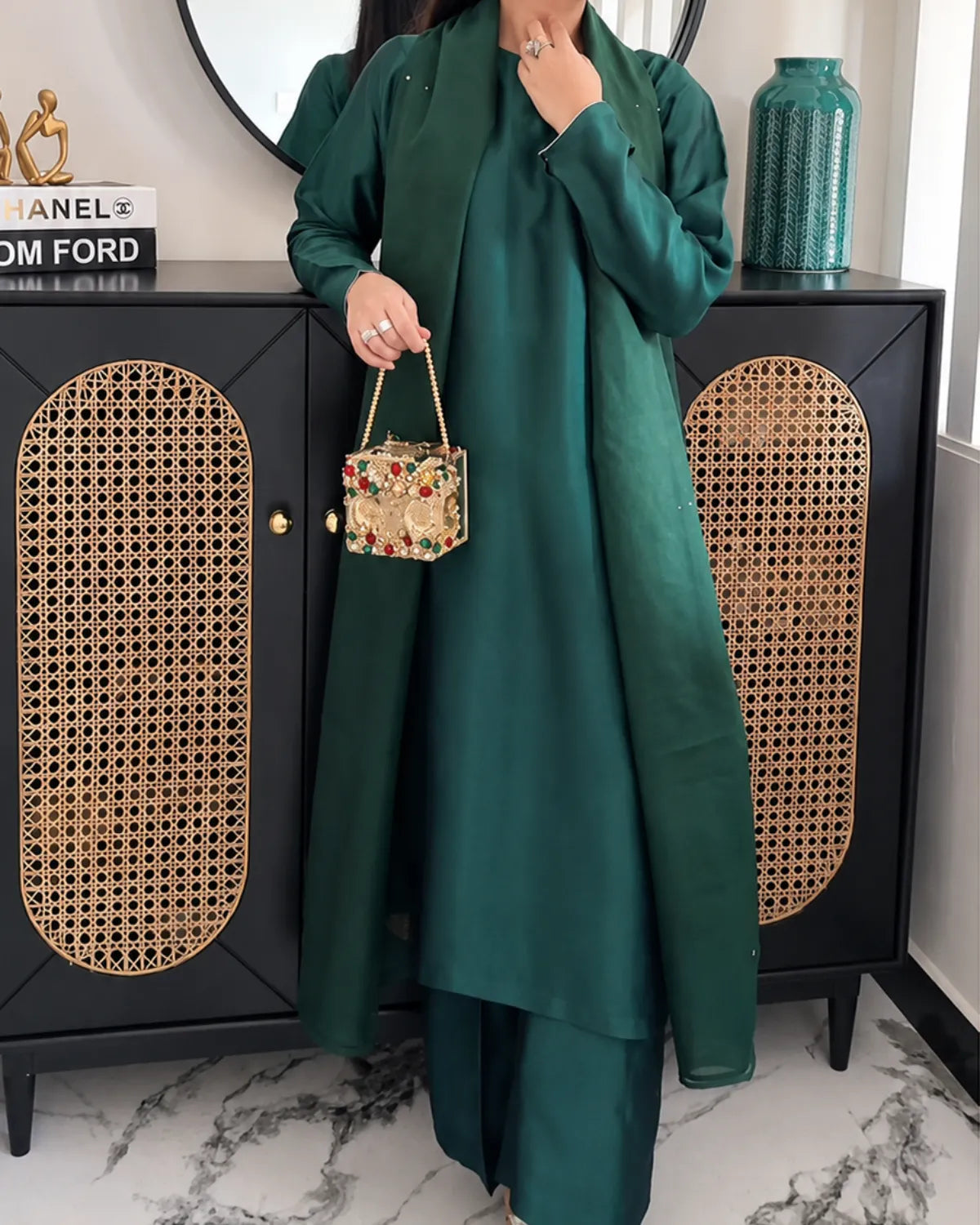 A woman models a dark green two-piece outfit consisting of a long-sleeve kurta and matching trousers, holding a small floral-patterned handbag. She stands in front of a black cabinet with woven rattan panels.