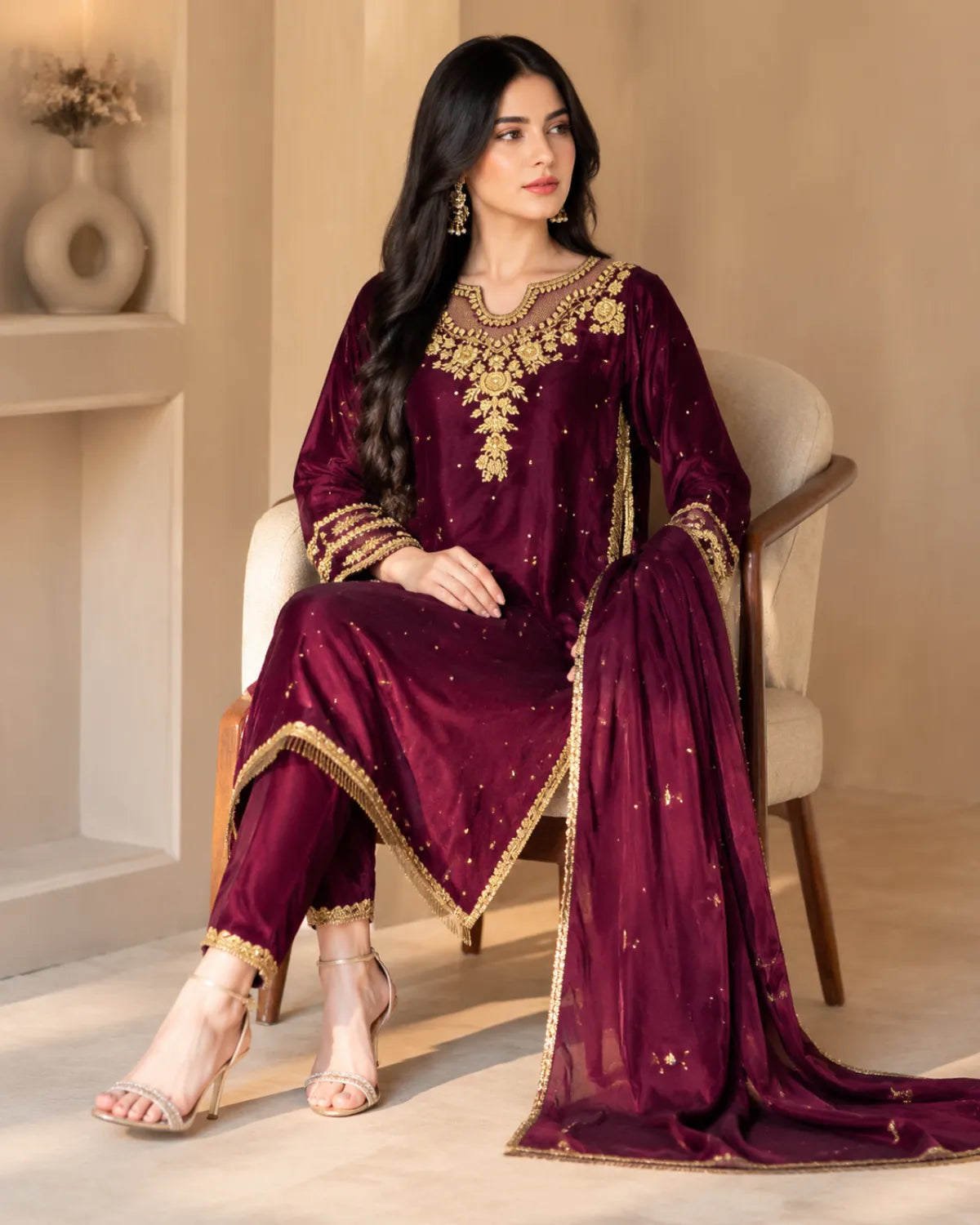 A woman seated in a cream armchair wearing a deep maroon ethnic outfit with ornate gold embroidery and a matching sheer dupatta, photographed in a warm neutral interior.