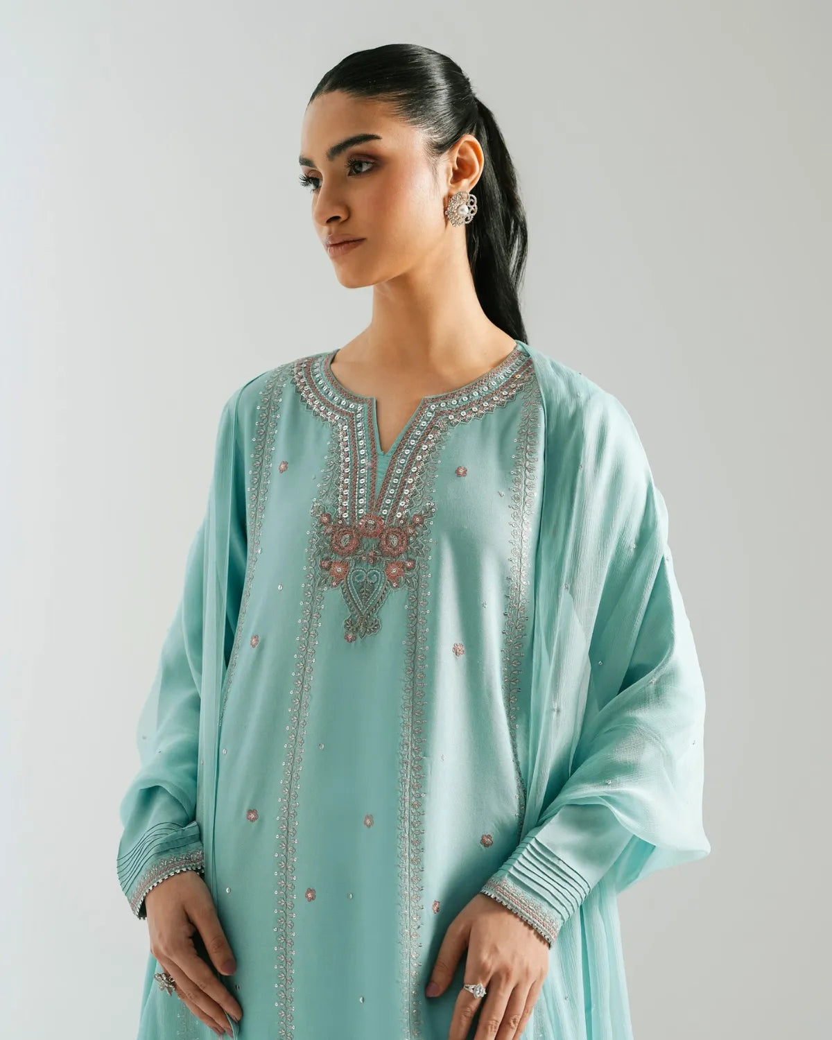 A woman wearing a mint green long-sleeve tunic with intricate silver and pink embroidery around the neckline, photographed against a neutral light gray background.