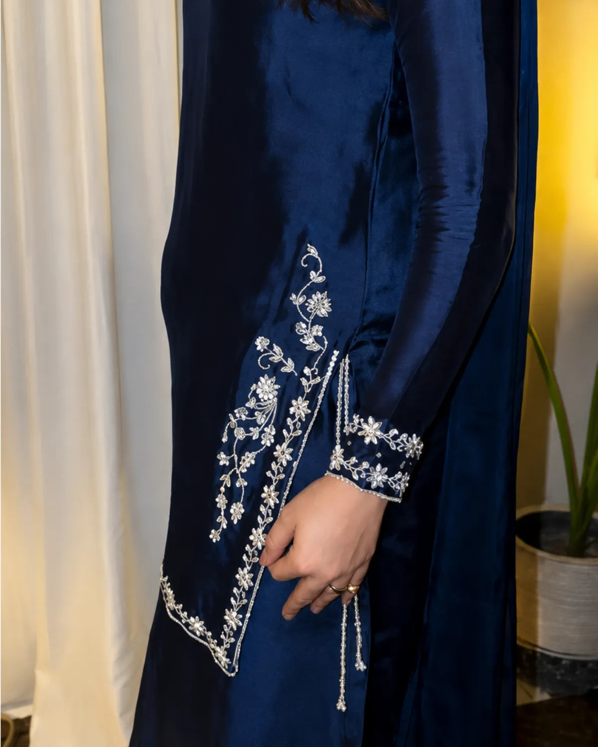 A close-up of a navy blue satin dress with intricate silver floral embroidery on the sleeve and hem, worn by a model in an indoor setting.