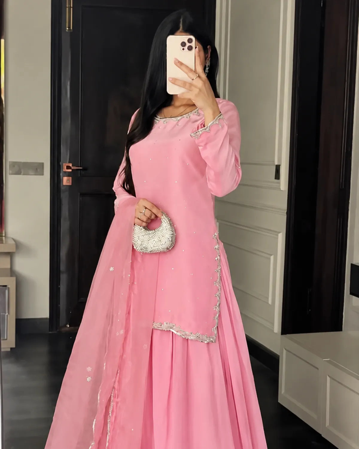 A woman takes a mirror selfie wearing a pink traditional long dress with silver embroidery and a matching sheer dupatta, holding a small silver purse.