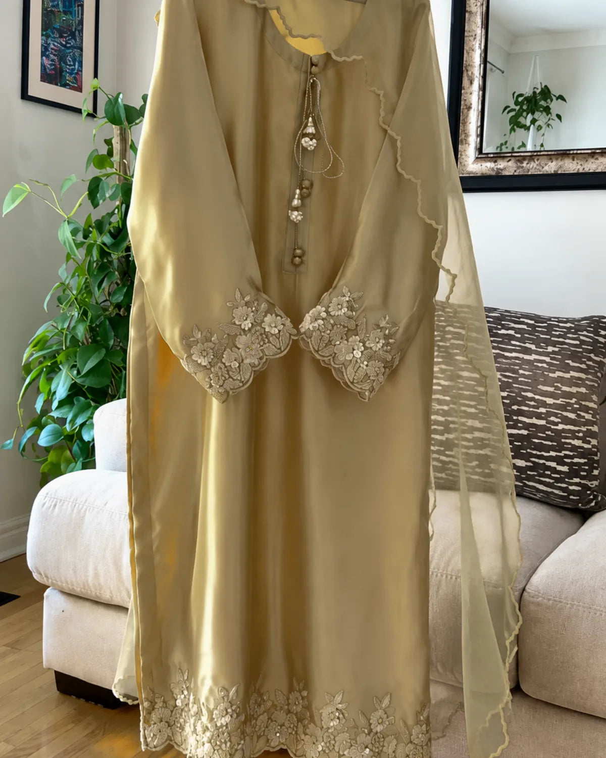 A beige satin-like long tunic or dress with floral embroidery on the cuffs and hem, hanging indoors in front of a sofa and mirror.