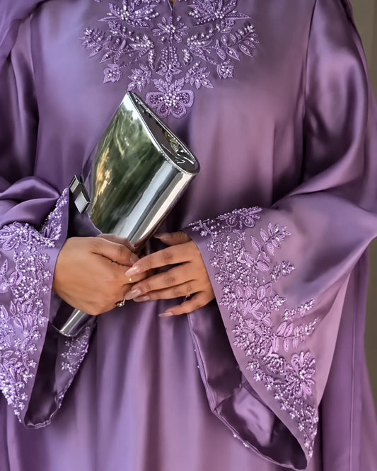 A close-up of a person wearing a lavender satin abaya with intricate floral embroidery and beadwork on the sleeves and neckline, holding a metallic silver clutch.