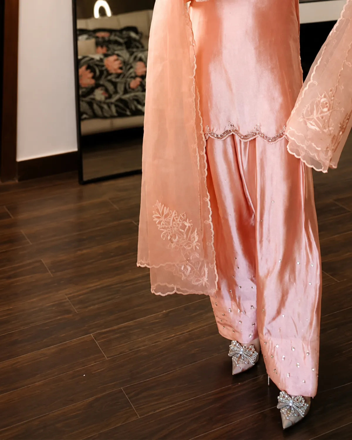 A person wearing a peach-pink traditional outfit with matching embroidered dupatta and wide-leg pants, standing on a dark wood floor in an indoor room.
