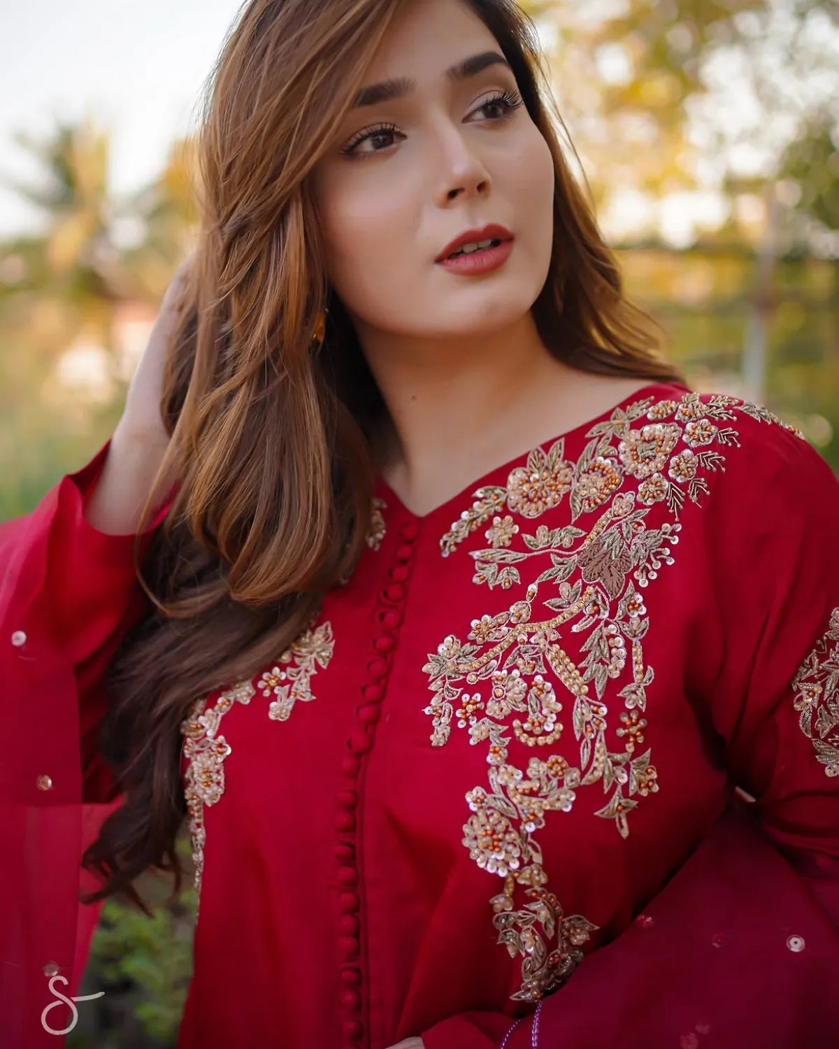 A woman wearing a deep red traditional embroidered outfit with intricate gold and beige floral embroidery on the neckline and sleeves, photographed outdoors with a soft blurred background.