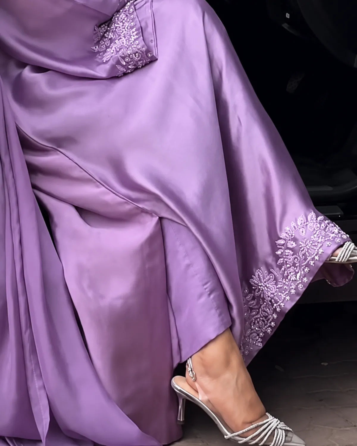 A close-up of a person wearing a flowing lavender satin abaya with ornate embroidered cuffs and a matching embroidered sleeve, paired with metallic heeled sandals.