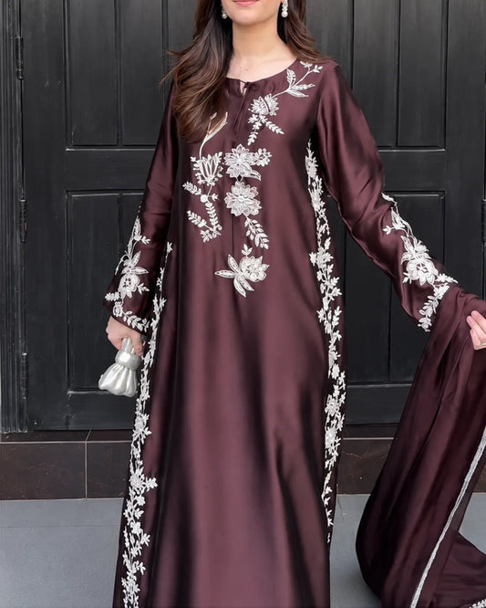 A woman wearing a long-sleeved, floor-length maroon dress with intricate white floral embroidery, holding a matching maroon dupatta in front of a dark paneled door.