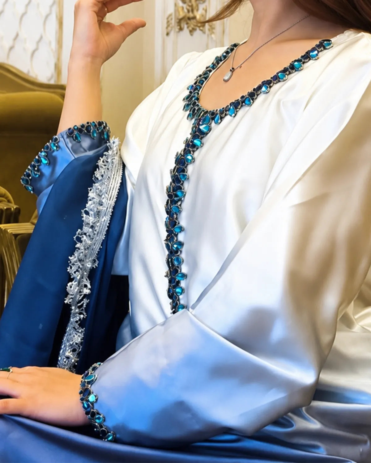 A close-up of a woman wearing a white satin robe with a deep V-neckline and wide sleeves, heavily embellished with sparkling blue and silver rhinestones. A matching blue sash or shawl with ornate silver embroidery and blue rhinestone trim is draped over her arm.