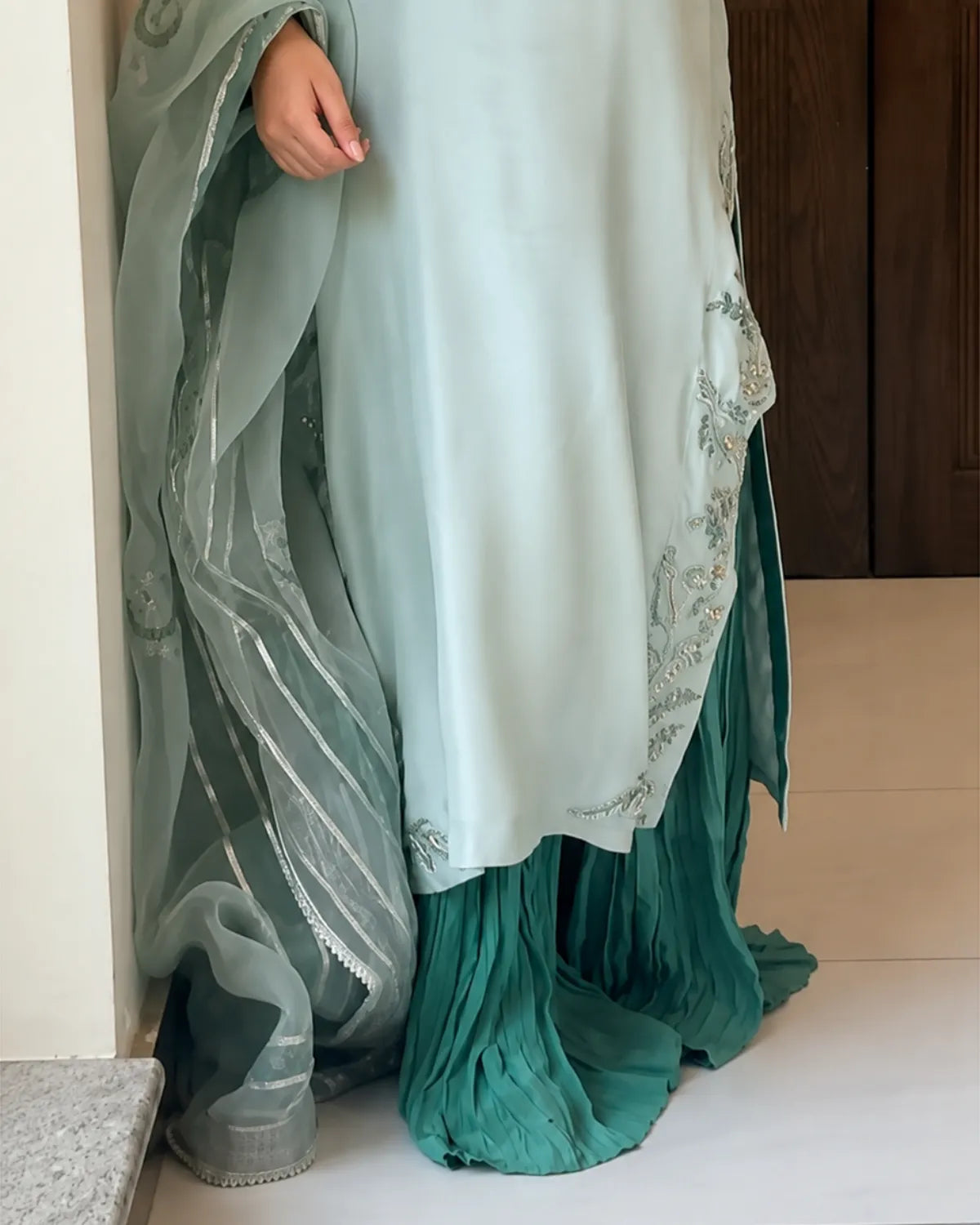 A close-up of a person wearing a traditional outfit with a light mint green top, a darker teal pleated skirt, and a matching sheer dupatta with silver embroidery, standing indoors.