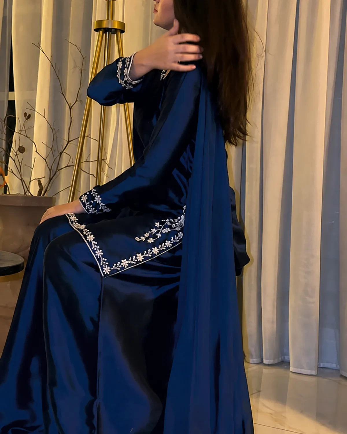 A woman wearing a long, flowing navy blue satin abaya with white floral embroidery on the cuffs and hem, seated in front of beige curtains.