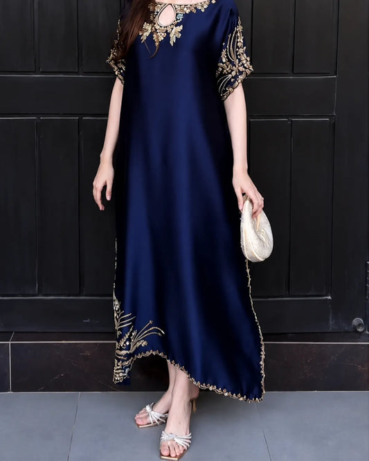 A woman wearing a navy blue maxi dress with gold embroidery on the neckline, sleeves, and hem, holding a small cream clutch against a dark paneled door background.