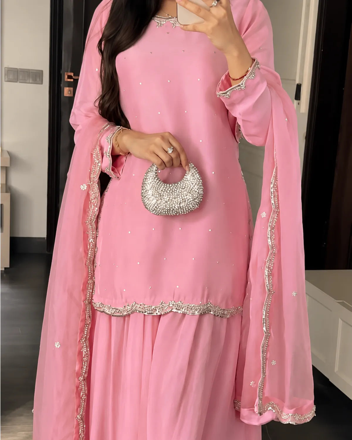 A woman wearing a pink traditional outfit with silver embellishments and a matching sheer dupatta, holding a small silver beaded handbag in an indoor setting.