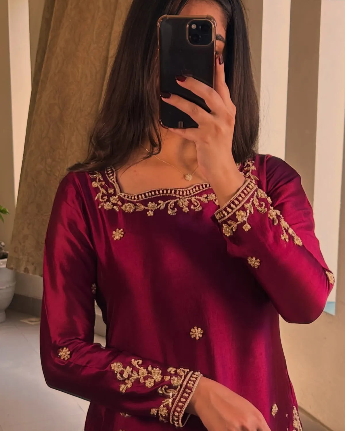A woman taking a mirror selfie wearing a long-sleeved magenta satin kurta with gold floral embroidery on the neckline and cuffs.