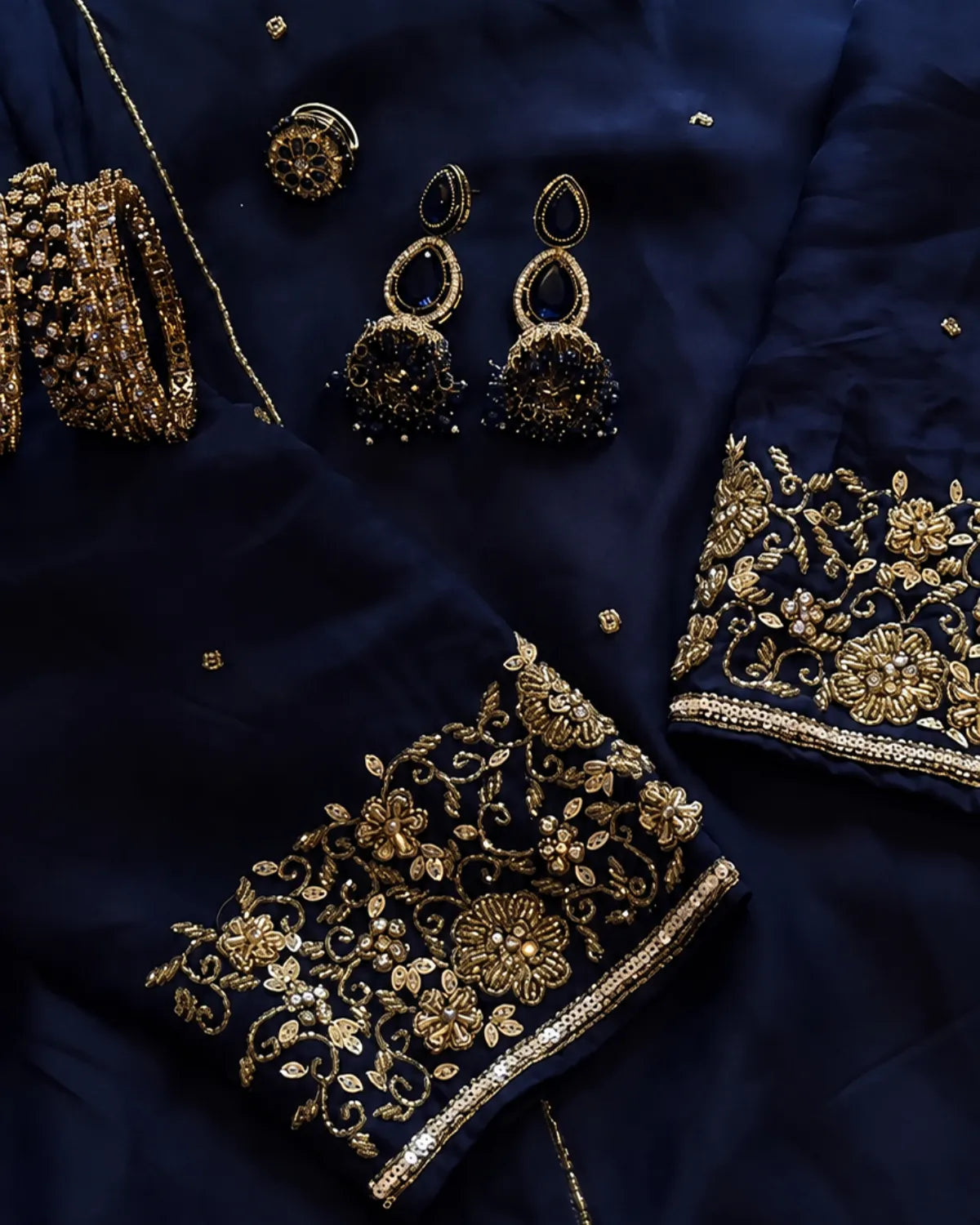 A close-up of a navy blue fabric garment with ornate gold floral embroidery and beadwork on the cuffs, alongside matching gold jewelry pieces including earrings and a bracelet, arranged on the fabric.