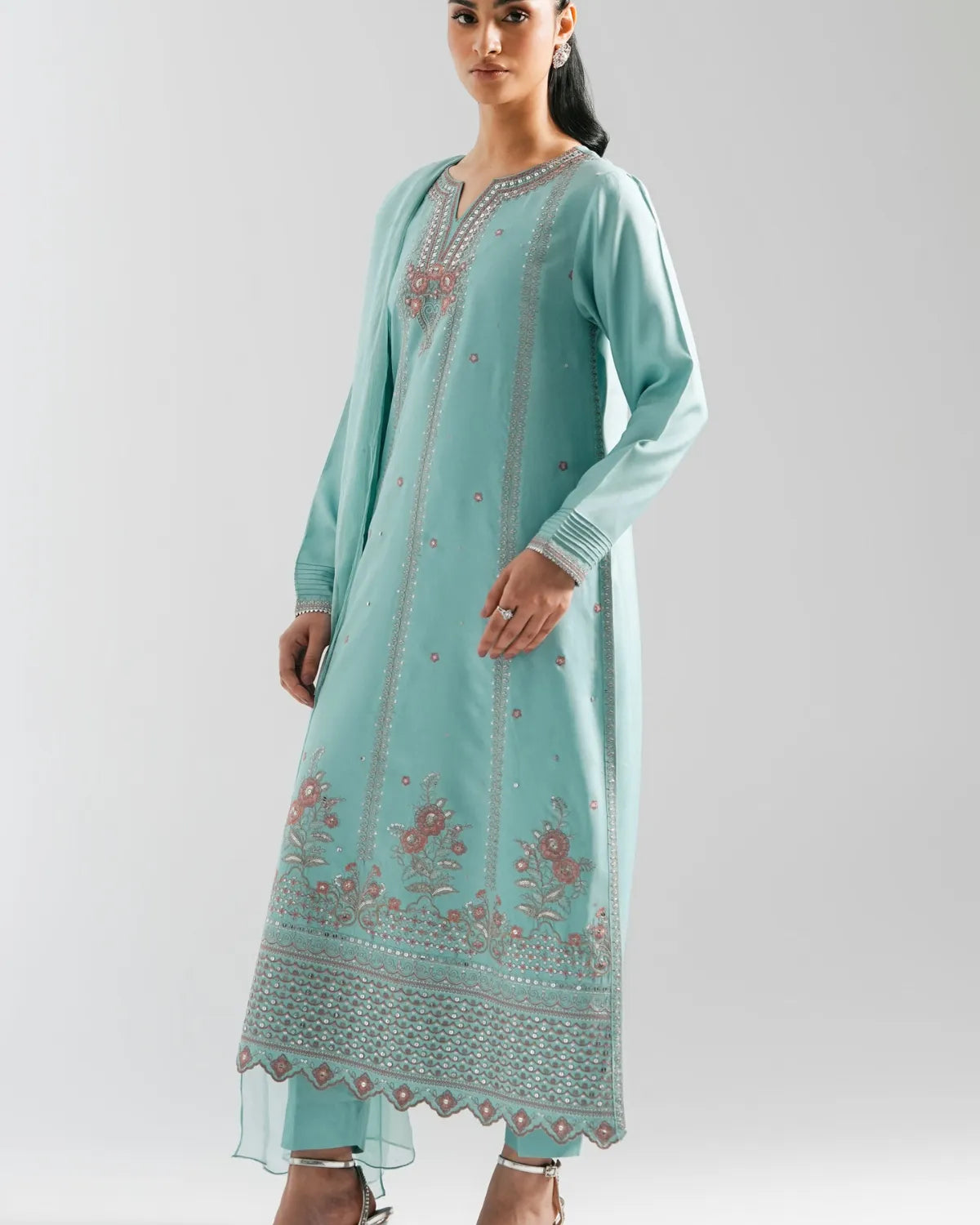 A woman modeling a long mint green kurta with pink floral embroidery and silver mirror work details, paired with matching trousers, against a light gray studio background.