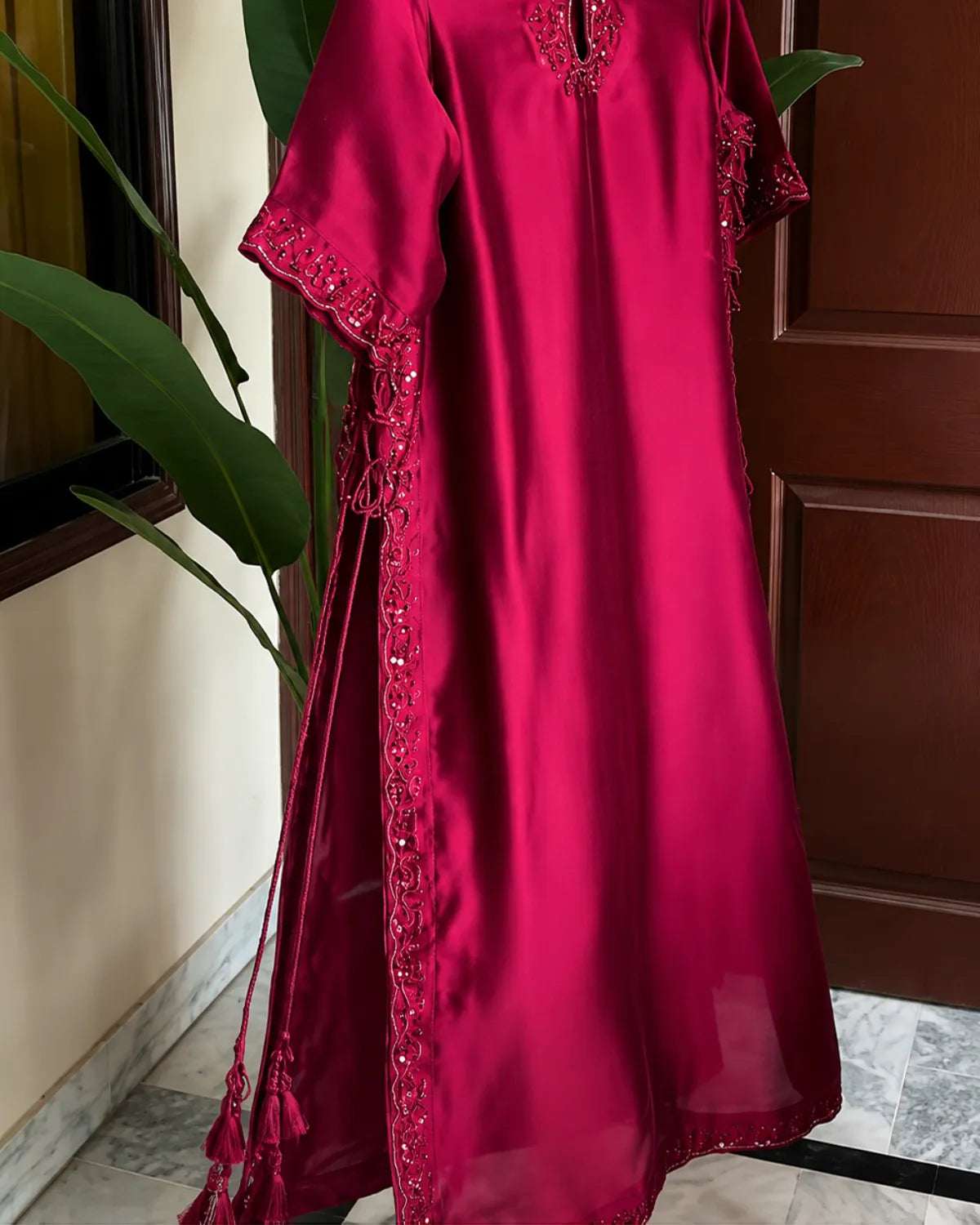 A bright magenta pink satin caftan dress with intricate embroidery and tassel details, hanging indoors next to a green plant.