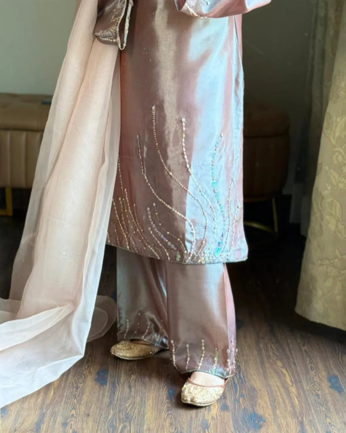 A person wearing a shiny blush pink traditional outfit with matching wide-leg pants and a sheer dupatta, standing on a wooden floor indoors.