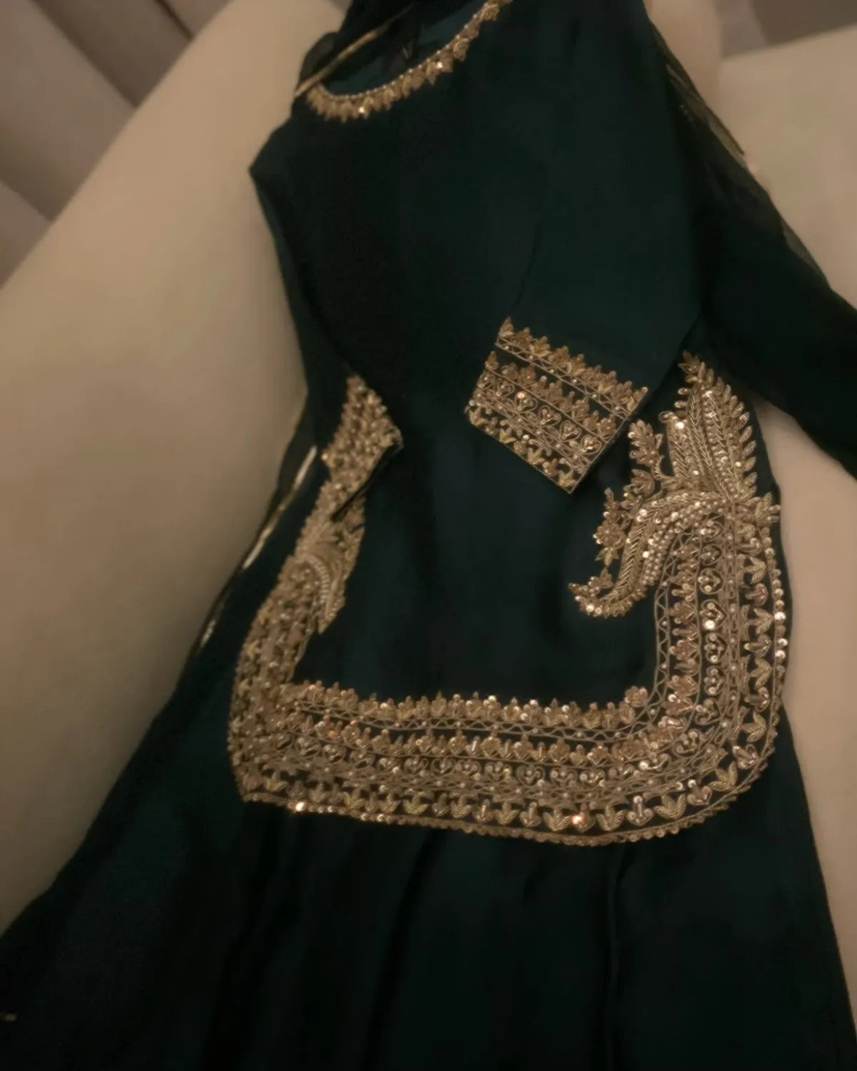 A dark green traditional dress with extensive gold and silver mirror work embroidery, shown draped on a light-colored sofa.