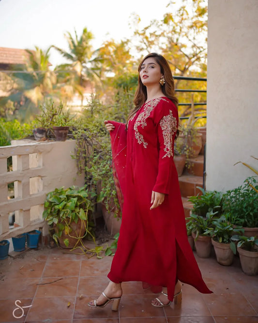 A woman wearing a long red embroidered dress stands on an outdoor patio with potted plants and warm sunlight in the background.