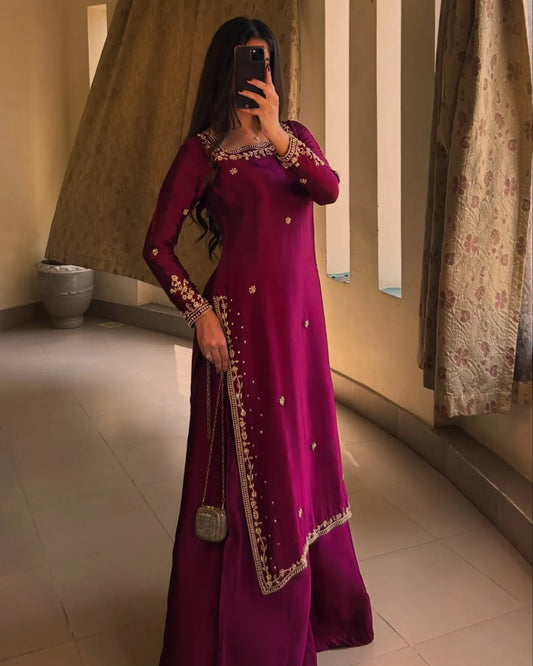 A woman takes a mirror selfie wearing a long magenta-purple traditional dress with gold embroidery and a matching skirt. She is holding a small metallic purse in a tiled indoor hallway with curtains.
