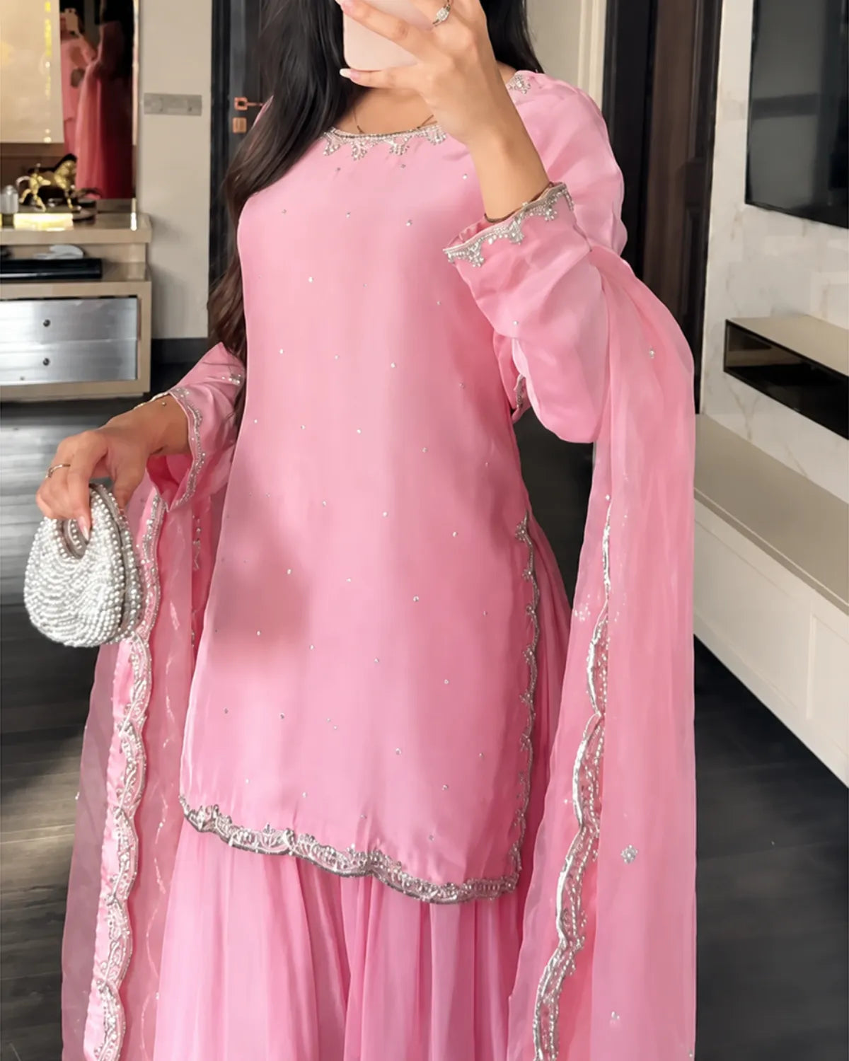 A woman wearing a pink traditional outfit with silver embroidered borders and a matching sheer dupatta, holding a small silver beaded clutch in an indoor room.