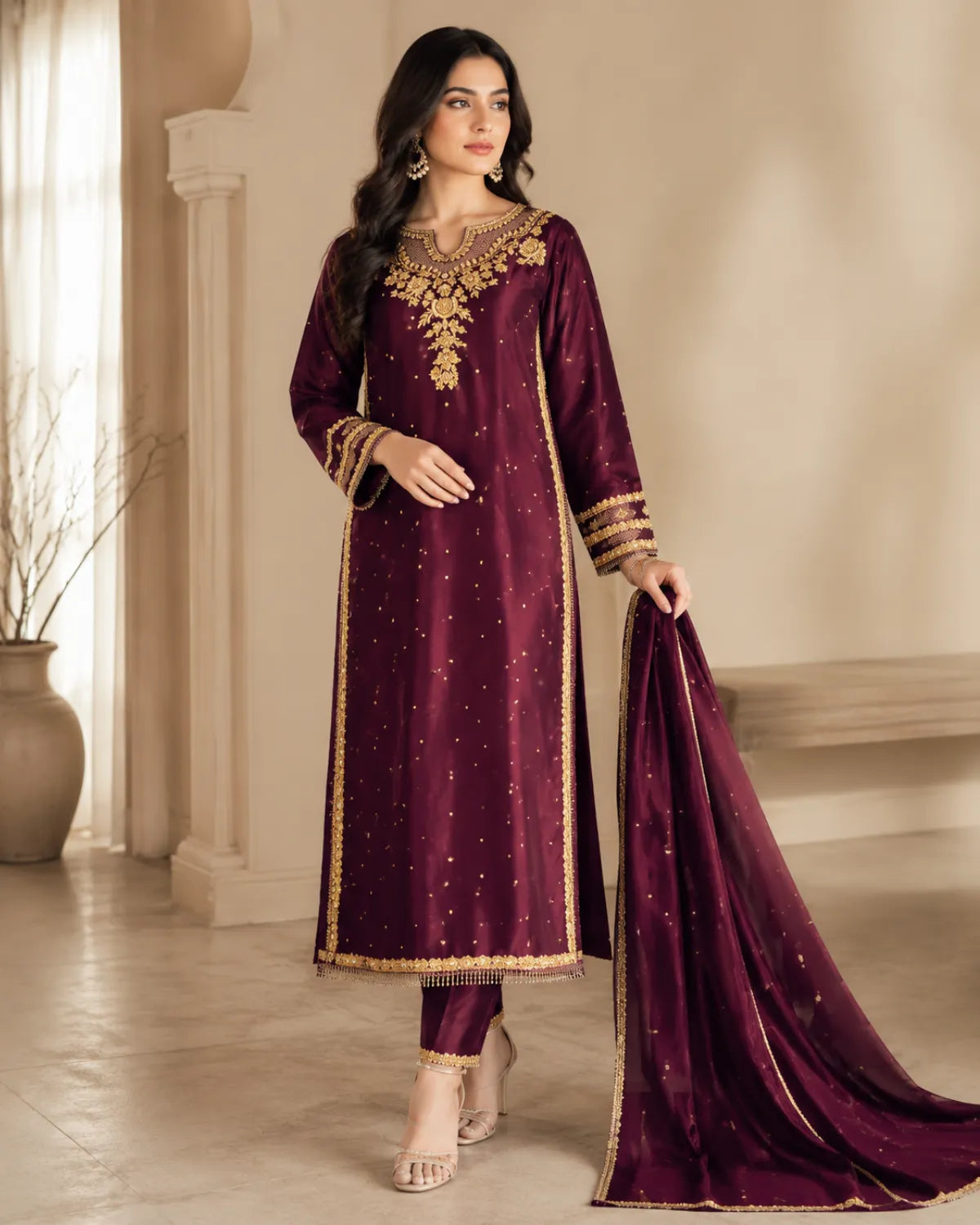 A woman models a deep maroon ethnic suit with gold embroidery and a matching dupatta in a warm, elegant indoor setting.